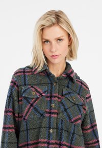 Groen geruite fleece shirtjas met blauwe en roze accenten, voorzien van een knoopsluiting aan de voorkant, twee borstzakken en een kraag. Zachte textuur.