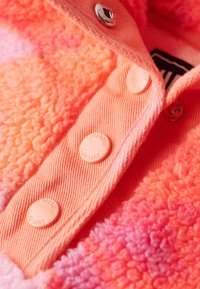 Blød fleecejakke i gradient lyserød og orange med en denim-lignende stolpelukning og prægnede knapper mærket "Superdry." Tekstureret stofdetalje synlig.