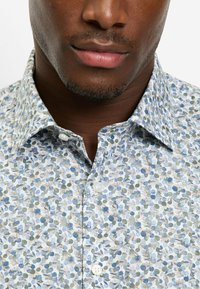 Blaues Hemd mit Blumendruck, versehen mit grünen und beigen Blattmustern, einem Kragen und weißen Knöpfen.
