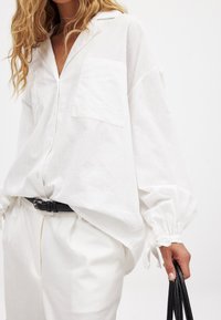 Balta ilga rankovė lininė marška su sagstomu priekiniu uždarymu ir krūtine kišenėje. Ypatybės - elastinės rankogaliai ir laisvas įsilaisvinimas. Suderinta su baltomis kelnėmis ir juodu diržu.