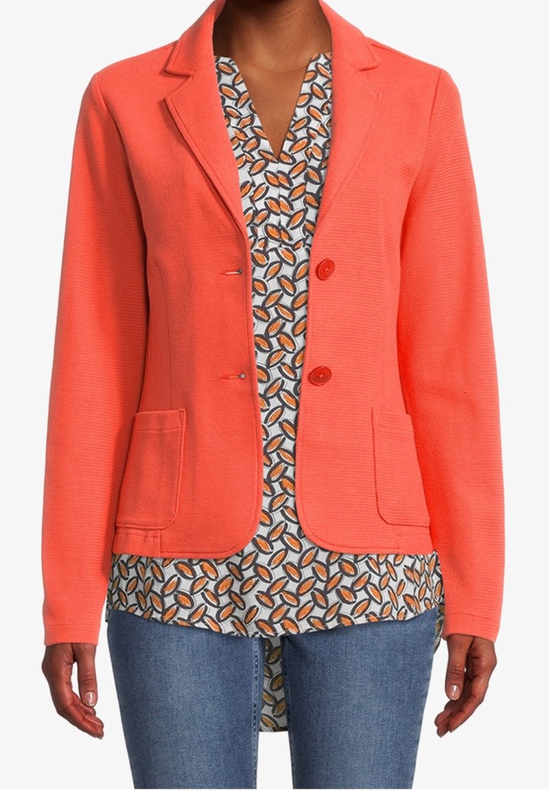 Femme portant un blazer corail sur un chemisier à motifs de feuilles et un jean bleu, debout devant un fond blanc uni.