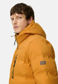 Orange polstret jakke med hætte, med en frontlynlås, justerbar snor og et logo patch på ærmet. Teksturomslag med isolering.