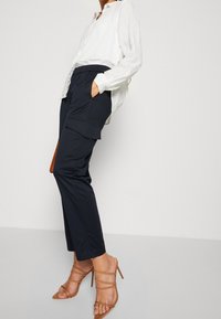 Witte lange mouwen knoopshirt, marineblauwe cargobroek met zijzakken, en tan gekruiste hoge hakken. Gladde stof met een ontspannen pasvorm.