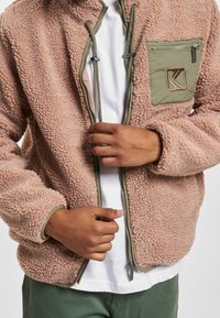 Polarowy sweter w delikatnym różu z zamkiem z przodu, zielonymi akcentami i kieszenią na piersi. Tekstura jest puszysta, w luźnym kroju i okrągłym kołnierzu.