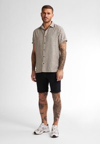 Camicia a maniche corte con bottoni, a righe verticali beige e marrone scuro, abbinata a pantaloni corti neri arrotolati e sneaker bianche.