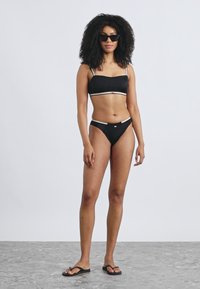 Melns bikini komplekts ar bantveida augšdaļu, kurai ir baltas akcenta daļas, un atbilstošām zemu jostasvietu apakšdaļām. Modelis to savieno ar melnām saulesbrillēm un šļūkšņām.