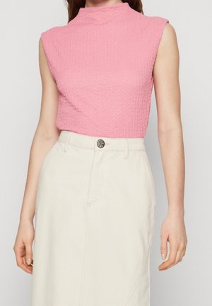 Femme portant un haut sans manches rose texturé rentré dans une jupe crème taille haute avec un bouton visible et des poches.