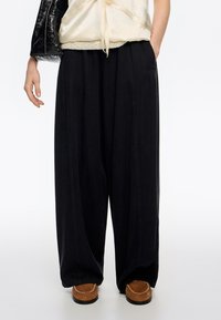 Czarne spodnie o szerokich nogawkach wykonane z gładkiej tkaniny, z bocznymi kieszeniami i ozdobnymi przeszyciami, zestawione z brązowymi butami z miękką podeszwą.