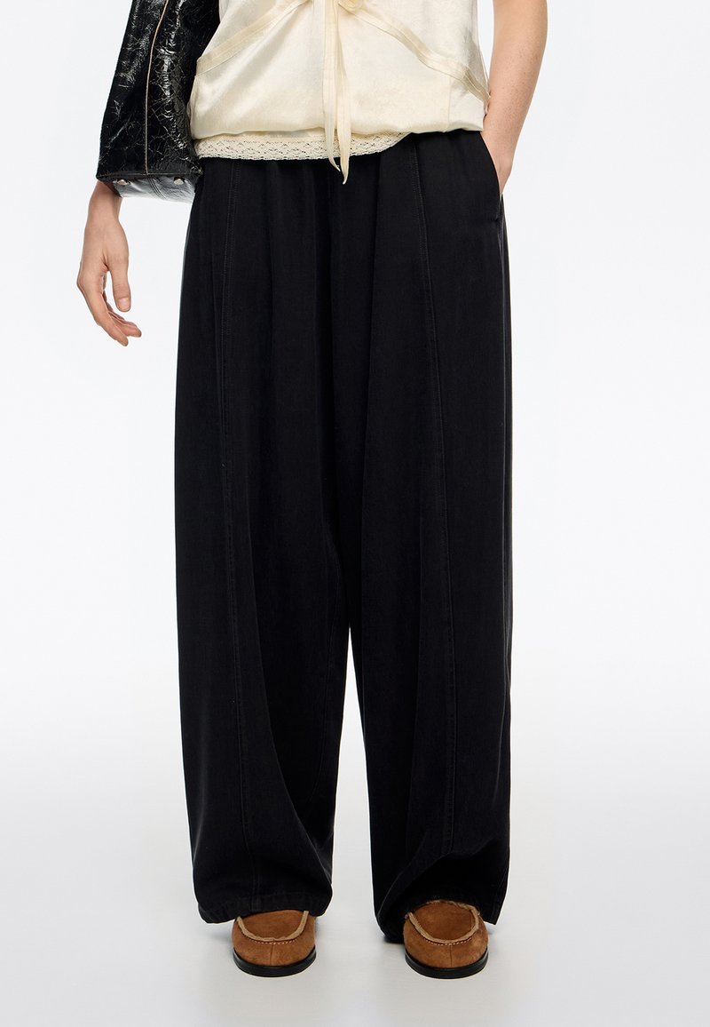 Czarne spodnie o szerokich nogawkach wykonane z gładkiej tkaniny, z bocznymi kieszeniami i ozdobnymi przeszyciami, zestawione z brązowymi butami z miękką podeszwą.