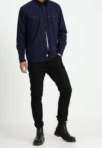 Námořnicky modrá košile s dlouhým rukávem a černým límcem, dvě přední kapsy, doplněná černými džíny a černými kotníkovými botami. Jednoduchý design a hladká textura.