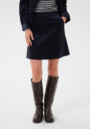 Femme portant une jupe en velours côtelé bleu marine, un haut rayé, une veste foncée et des bottes en cuir brun foncé jusqu'aux genoux, debout avec une main dans la poche.
