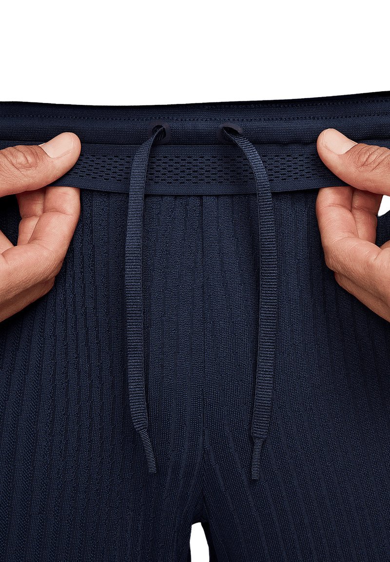 Pantalones de chándal azul marino con cintura ajustable por cordón, con tejido texturizado y un borde superior de punto plano. Manos acarician la zona de la cintura.