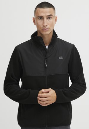 Schwarze Fleecejacke mit glattem oberen Bereich, hohem Kragen und durchgehendem Reißverschluss. Ausgestattet mit Seitentaschen und einem kleinen Logo auf der Brust.