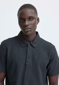 Donkergrijs poloshirt met een gestructureerde stof, korte mouwen en een klassieke kraag. Voorzien van drie knopen bij de hals.