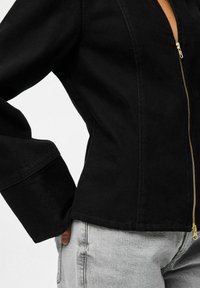 Črna jeans jakna z zadrgo spredaj, z dolgimi rokavi in prilegajočim krojem. Zlata zadrga dodaja kontrastno podrobnost.