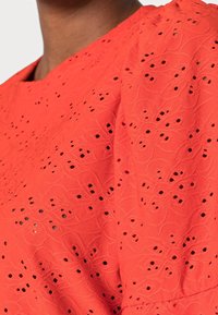 Tissu rouge brodé avec des motifs floraux découpés et une texture relief. Présente un col arrondi décontracté et des manches bouffantes.