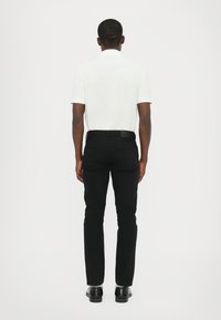 Cămașă polo albă, blugi negri, pantofi negri. Croială slim fit, mâneci scurte și culoare solidă. Vedere din spate care arată detalii despre cusături și buzunare.