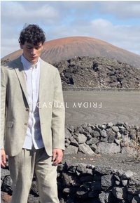 Ein Mann in einem hellbeigen Anzug und weißem Hemd steht nahe Vulkanfelsen, im Hintergrund ein großer Vulkankegel unter einem bewölkten Himmel.