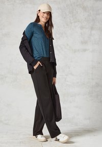 Lässiges Outfit mit einem blauen gemusterten Oberteil, schwarzer Jacke, schwarzen Hosen und weißen Sneakern. Eine beige Mütze setzt einen Akzent.
