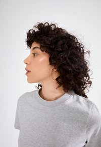 Pelēka kokvilnas T-krekls ar apaļu kakla izgriezumu; modelis ar cirtainiem tumšiem matiem un valkā zelta riņķa auskars; profila skats.