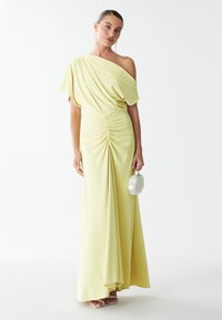 Robe jaune à une épaule avec un tissu drapé et un plissage sur le côté. Un mannequin tient un petit sac blanc, portant des talons beiges ouverts.