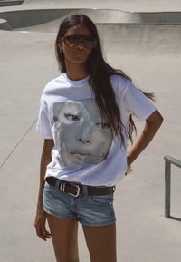 Hvid t-shirt med et stort monochromt ansigtsprint, parret med denimshorts og et sort bælte. Modellen bærer oversized solbriller.