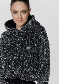 Czarna i srebrna kurtka z kapturem, charakteryzująca się teksturowanym, cekinowym wzorem. Zawiera czarny guzik z detalem nuty muzycznej.