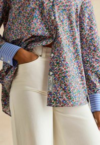 Floral-patterned shirt with multicolored flowers on a white background, featuring a blue striped cuff and a labeled hem. Paired with white pants.
