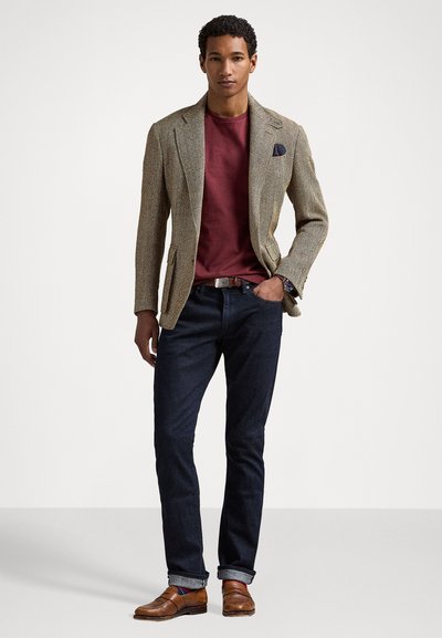 Blazer en chevrons marron sur une chemise bordeaux, accompagné d'un jean foncé et de chaussures en cuir marron. Les détails notables incluent un pochette et des poignets retroussés.