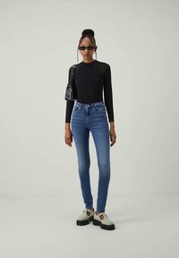 Mulher em pé, vestindo uma blusa preta de manga longa, jeans skinny azuis, sapatos brancos de plataforma, uma bolsa preta de ombro e óculos de sol pequenos e pretos.
