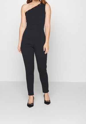 Czarny kombinezon na jedno ramię z koronkową lamówką, dopasowaną talią i zwężanymi nogawkami. Wykonany z teksturowanej tkaniny, w połączeniu z czarnymi butami na obcasie.