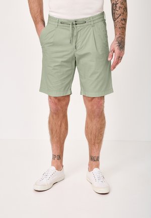 Man wearing light green drawstring shorts, white sneakers, and a white shirt, with tattoos on legs and right arm visible.
