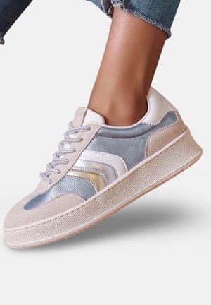 Pied portant une basket basse avec des panneaux en daim beige clair et gris, des accents blancs et métalliques, et une semelle épaisse texturée, associée à un jean retroussé.