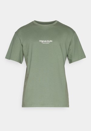 T-shirt vert olive en coton doux, doté d'un col rond et de manches courtes. Il comporte un texte blanc indiquant "Originals Studio" sur le devant.