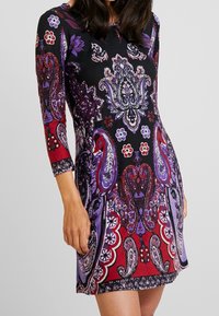 Femme portant une robe noire ajustée avec des motifs floraux et cachemire complexes en violet, rouge et blanc, avec des manches trois-quarts.