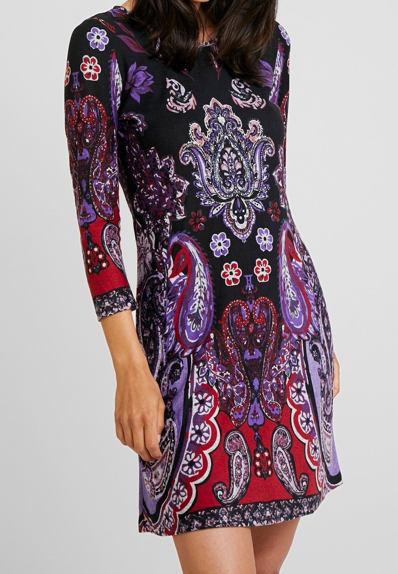 Femme portant une robe noire ajustée avec des motifs floraux et cachemire complexes en violet, rouge et blanc, avec des manches trois-quarts.