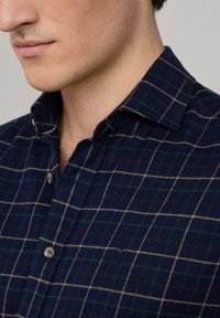 Camicia di flanella blu navy con motivo a scacchi verde e beige, colletto con bottoni e accento del logo discreto. Il tessuto texturizzato suggerisce calore.