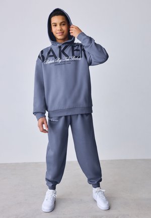 Adolescent portant un sweat à capuche gris et un pantalon de survêtement avec des baskets blanches, debout, ajustant sa capuche sur un fond uni.