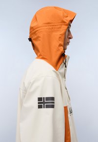 Chaqueta impermeable con un diseño bicolor, con capucha naranja y cuerpo crema. Incluye un logo negro en la manga.