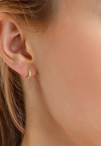 Zlatna omča s naušnicom u zakrivljenom, minimalističkom dizajnu. Glatka, sjajna površina s elegantnim profilom. Nosena na režnju uha.