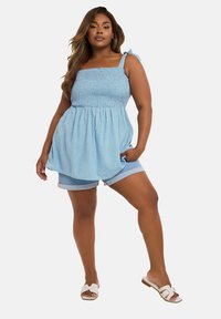 Hellblaues, schulterfreies Oberteil mit gerafftem Mieder und Bindebändern, kombiniert mit Denim-Shorts. Der Stoff hat kleine weiße Punkte.