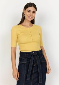 Gul ribbad kortärmad topp med rund halsringning och framknappar, kombinerad med en mörk denimkjol med knytband i midjan och fickor.