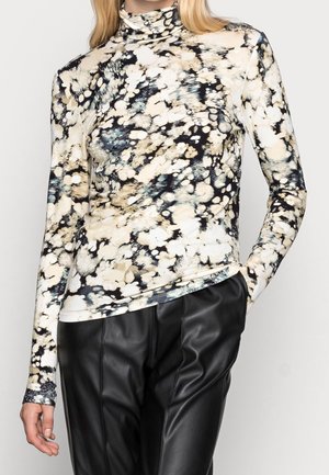 Kvinde iført en langærmet, højhalset top med abstrakt print i sort, beige og hvid, sat sammen med sorte læderbukser.