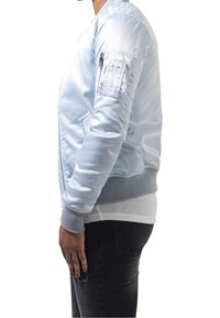 Błękitna satynowa kurtka bomberka z prążkowanymi mankietami, opływowym kształtem oraz kieszenią na suwak na lewym rękawie, noszona na białej koszuli.
