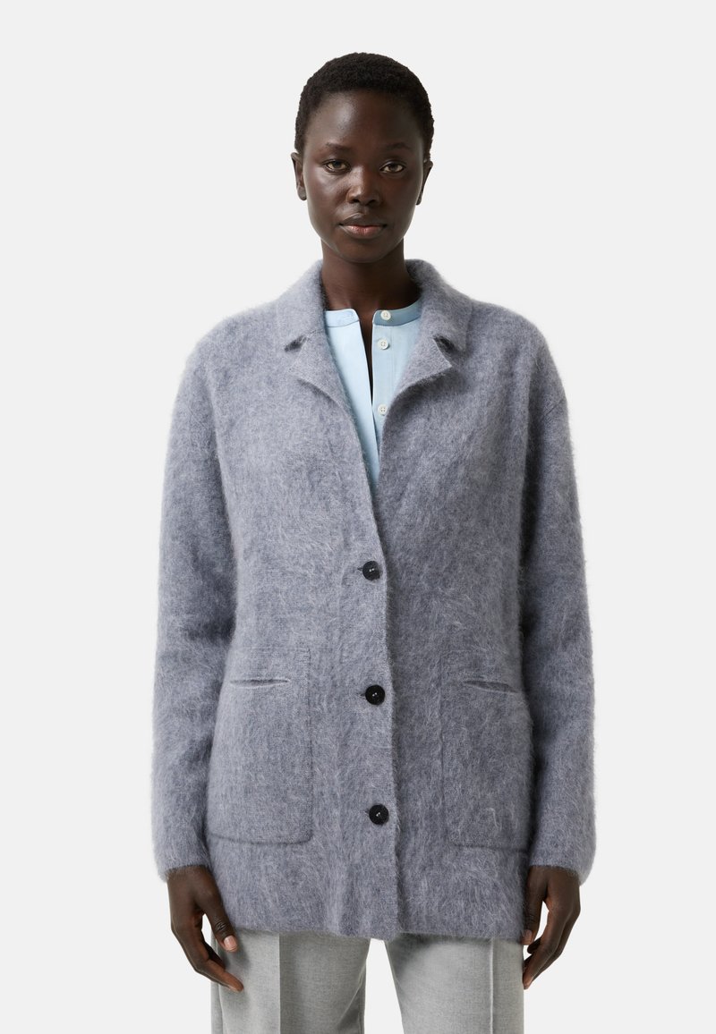 Woman wearing a soft, fuzzy light gray buttoned cardigan over a light blue shirt and gray pants, standing against a plain background.