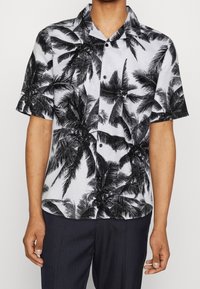Chemise à manches courtes boutonnée en blanc avec un motif de palmiers noirs. Tissu léger et respirant avec un col classique et des boutons sur le devant.