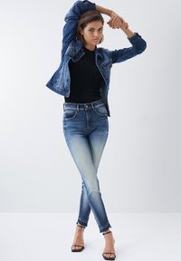Chaqueta de mezclilla con un lavado azul clásico, combinada con una camiseta negra de canalé y jeans skinny de tiro alto. Presenta un dobladillo deshilachado y diseño de apertura frontal.