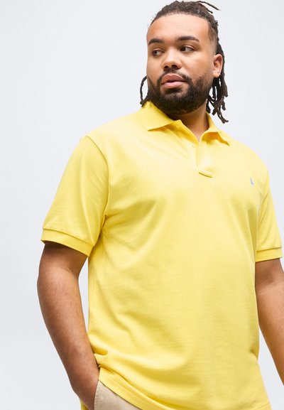 Άνδρας με γένια και ντραντλοκς που φοράει κίτρινο πόλο με μικρό λογότυπο, κοιτάζει στο πλάι με το ένα χέρι στην τσέπη των μπεζ παντελονιών.