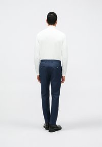 Biela dlhá blúzka s dlhým rukávom v kombinácii s tmavomodrými priliehavými nohavicami s dvoma zadnými vreckami a hladkou textúrou, nosené s čiernymi topánkami.