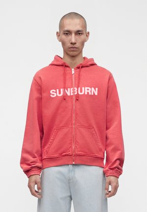 Mand med barberet hoved iført rød hættetrøje med lynlås med teksten "SUNBURN" og lyseblå jeans, stående mod en enkel hvid baggrund.